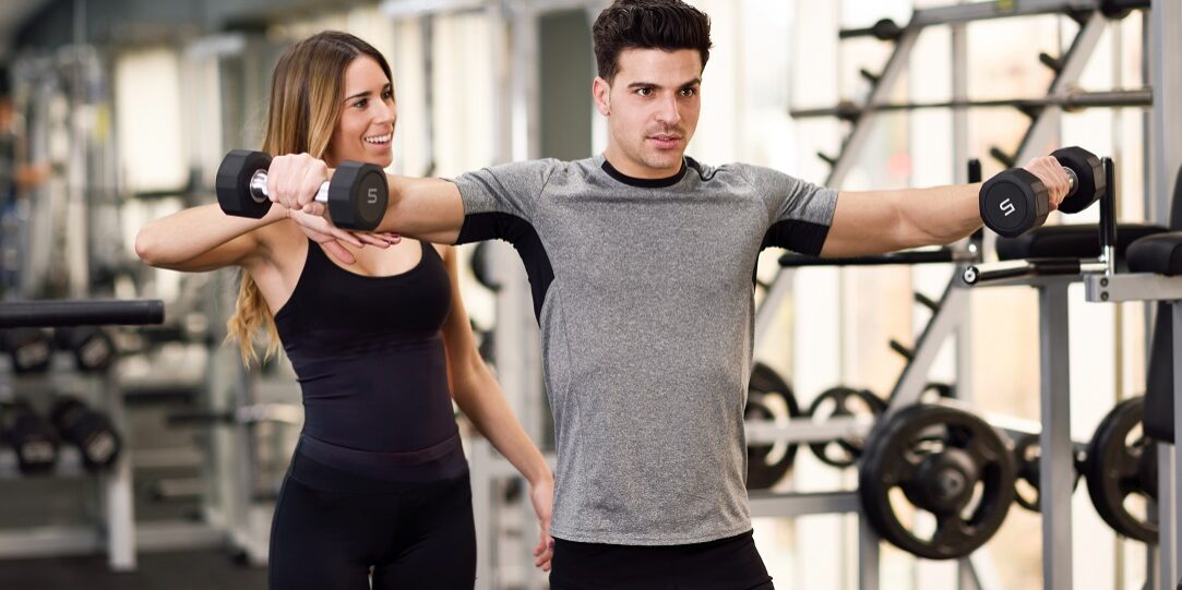 Eine Personal Trainerin korrigiert die Übung eines Kunden beim Seitheben mit Kurzhanteln im Fitorama Basel durch gezielte Berührung am Arm.